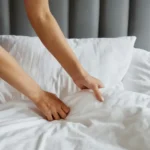 Mulher arrumando e organizando lençóis brancos na cama com cabeceira cinza