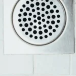Ralo de banheiro limpo com tampa metálica sobre piso de cerâmica branca, representando solução para mau cheiro no ralo do banheiro.