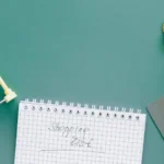 Um carrinho de compras em miniatura, um caderno com a inscrição "Shopping List", um cartão de crédito e uma pequena sacola de papel, todos dispostos sobre um fundo verde-azulado.